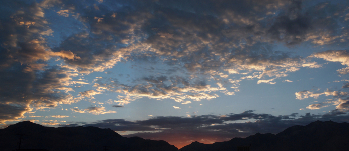 [Several photos stitched together to give a wide view of the mountains with a small triangular tinge of orange red in the mountain pass area. Obove it are light blue skies with a series of small white clouds high above.]